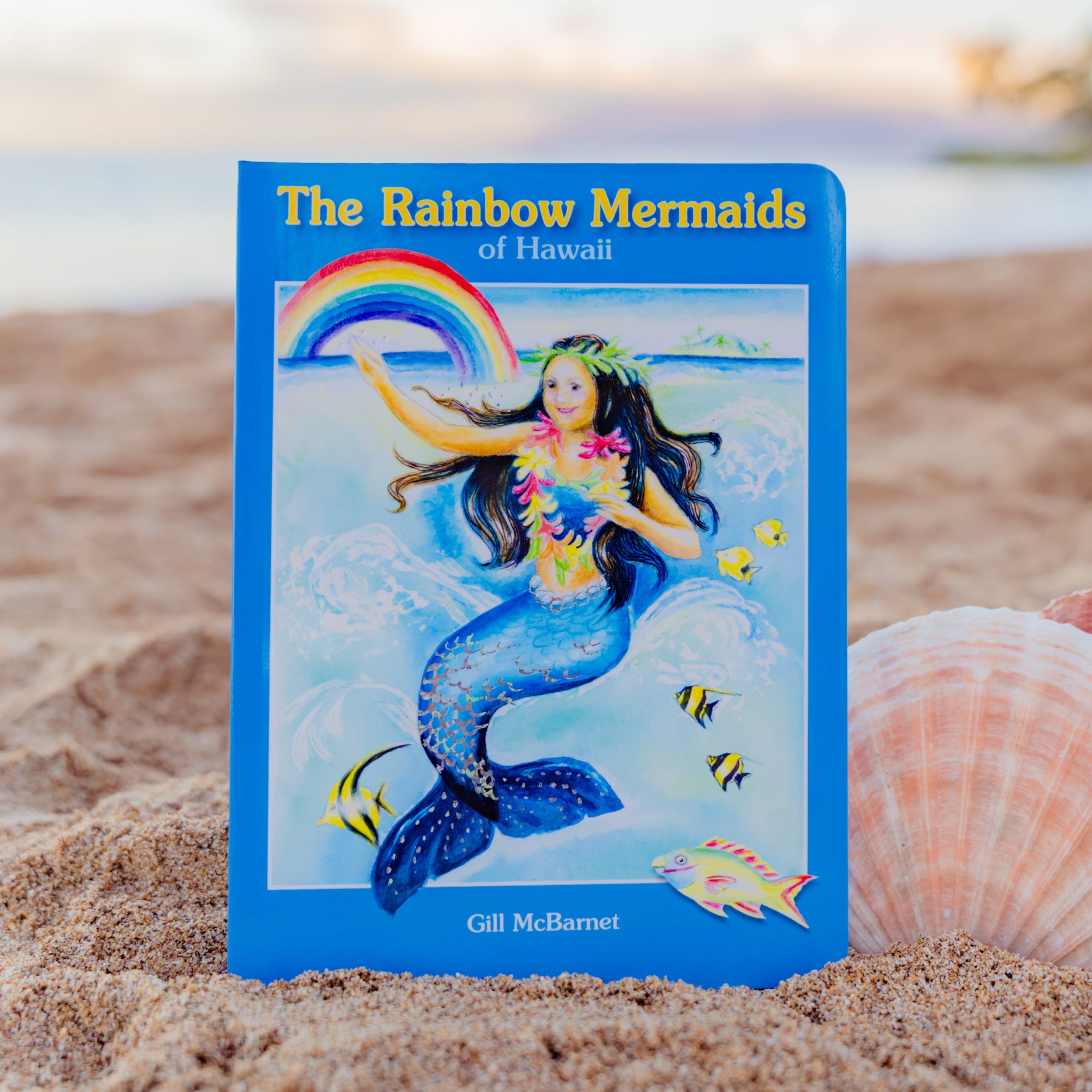 Book titled 'The Rainbow Mermaids of Hawaii' on a sandy beach with shells.